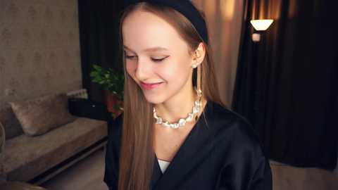 Media: Video of a young woman with straight brown hair, wearing a black robe and pearl necklace, smiling softly. Background features a beige couch, green plant, and dark curtains.