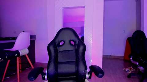 Media: Video of a black gaming chair in a dimly lit room with purple lighting, featuring a white chair and desk to the left, and cluttered floor space to the right.