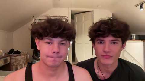 Media: Video of two young men with curly hair, one with a black tank top, the other in a black shirt, standing in a cluttered attic bedroom with white walls and a door.