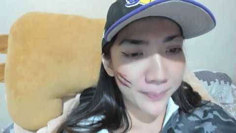 Media: Video of an Asian woman with long black hair, wearing a black cap with yellow logo, lying on a bed with a plush teddy bear. Her face has a small cut on her cheek.