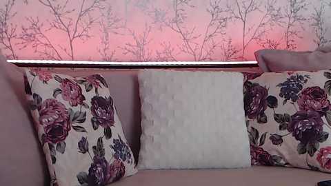 Media: A video of a cozy living room with a pastel pink floral wallpaper and a plush cream-colored sofa adorned with decorative pillows featuring large, vibrant floral patterns in shades of purple and green.