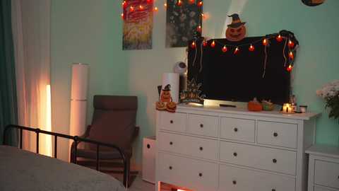 Media: A video of a cozy, Halloween-themed bedroom with a white dresser adorned with jack-o'-lanterns, a black TV, string lights, and a witch hat. A gray bed and chair are visible, and the walls are painted mint green.