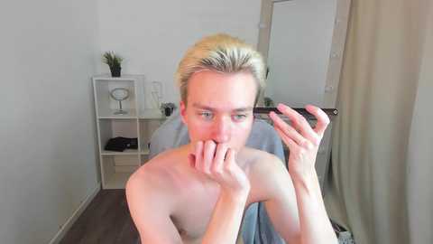 Media: Video of a topless, fair-skinned man with short, platinum blonde hair, holding a lipstick, in a sparsely furnished room with white walls, a white shelf, and a mirror.