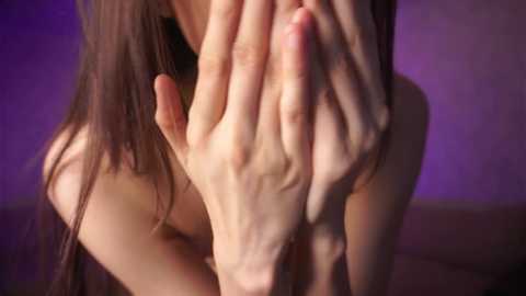 Media: Video of a woman with long brown hair covering her face, hands clasped in front. She appears to be topless, with a blurred purple background suggesting an indoor setting.