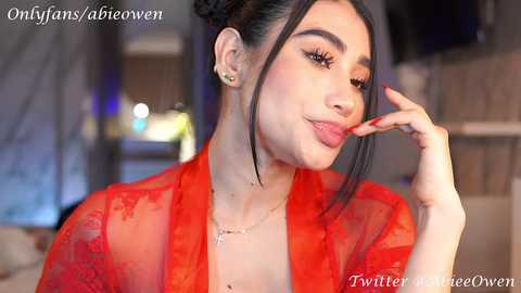 Media: Video of a Latina woman with light skin, black hair styled in a bun, wearing a sheer red lace robe. She has a confident, playful expression with red lipstick and manicured nails.