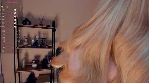 Media: Video of a person with long, wavy blonde hair, captured from the side. Background features a beige wall and a shelf with various items, including a plant, bottles, and decorative objects.