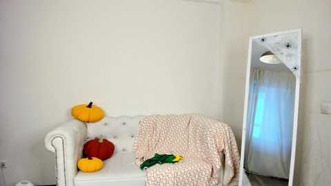 Media: Video of a minimalist, cozy living room with a white tufted sofa draped in a patterned blanket, adorned with three pumpkins in red, yellow, and orange, a green scarf, and a white mirror with star-shaped cutouts.