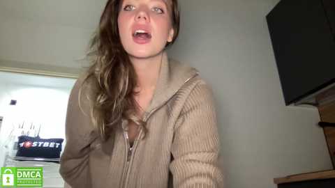 Media: Video of a young woman with long, wavy brown hair, wearing a beige zip-up hoodie, looking surprised or excited, indoors with a TV and kitchen visible in the background.