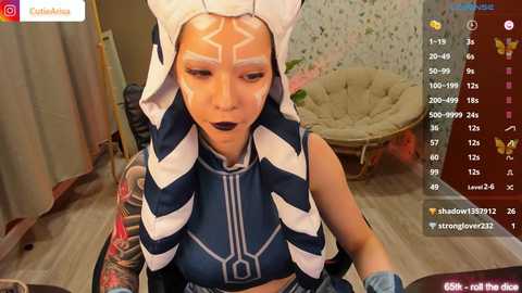 Media: Video of a woman wearing a Star Wars-inspired costume, featuring Ahsoka Tano's headpiece and makeup. She sits on a chair in a cozy room with floral wallpaper and a beige chair. Streaming platform interface with chat and viewer count visible.