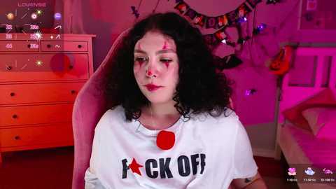 Media: Video of a young woman with curly black hair and a white T-shirt with \"FUCK OFF\" in bold letters, in a bedroom with pink lighting, a dresser, and a bed.