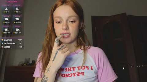 Media: Video of a young woman with long red hair, pale skin, and a tattooed arm, wearing a pink and white T-shirt, brushing her teeth in a dimly lit room.