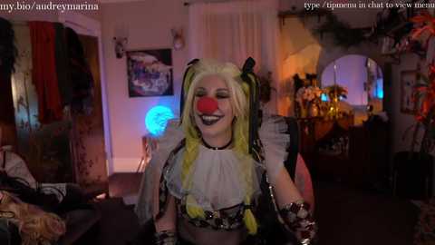 Media: Video of a woman in a colorful, intricate cosplay outfit, including a yellow wig, red nose, and black-and-white striped bodice, standing in a dimly lit, cluttered room with a festive background.