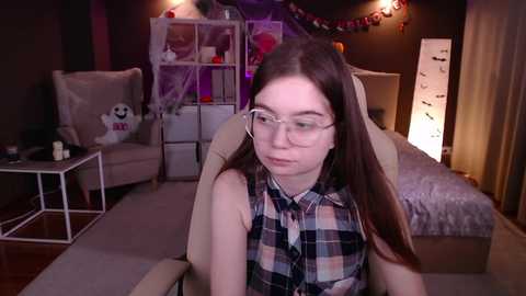 Media: Video of a young Caucasian woman with long brown hair and glasses, wearing a plaid shirt, sitting in a beige chair in a dimly lit bedroom with Halloween decorations.