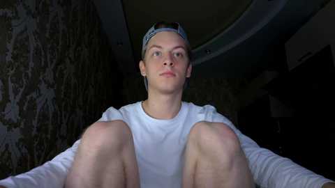 Media: Video of a young Caucasian man with short blonde hair, wearing a white t-shirt and a backwards cap, sitting on a couch with his knees up, in a dimly lit room with camo-patterned wallpaper and a curved ceiling.