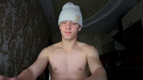 Media: Video of a young, shirtless, fair-skinned Caucasian male with short hair, wearing a white beanie with a small emblem, standing indoors with a patterned wallpaper and dim lighting.