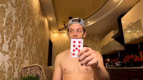 Media: Video of a shirtless young man with a light complexion, wearing a backward baseball cap, holding a playing card with red hearts over his face in a dimly lit, patterned wallpapered room.