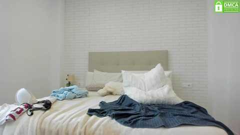 Media: Video of a minimalist bedroom with white brick wall, beige headboard, unmade bed with white sheets, blue blanket, and scattered clothes, including a white hat and black shoes.