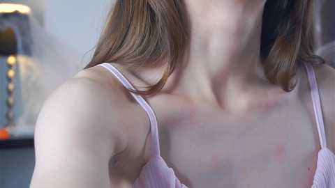 Media: Video of a woman's upper torso in a light pink, ribbed tank top, showing slight redness on her neck and shoulders, blurred background with a lamp and a dark surface.