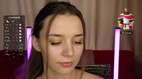 Media: Video of a young woman with light skin and brown hair, wearing minimal makeup, seated in a room with a digital clock, a gold statue, and a red sofa.