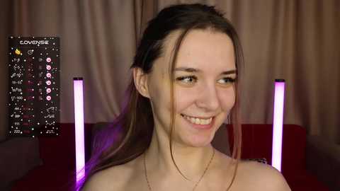 Media: Video of a smiling young woman with light skin, brown hair, and a slender build, wearing a thin gold necklace, against a backdrop of red curtains with purple LED lights.