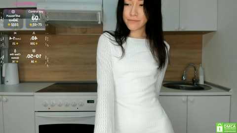 Media: Video of a slender, East Asian woman with long black hair, wearing a white knitted sweater, standing in a modern kitchen with a white stove, wooden backsplash, and a silver faucet.