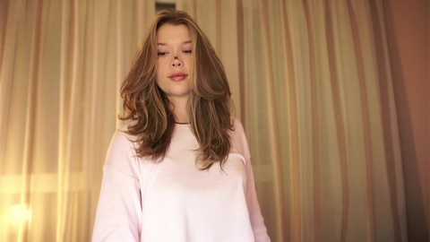 Media: Video of a young woman with long, wavy brown hair, wearing a loose white sweater, standing in front of a softly lit, sheer curtain background.