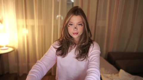 Media: Video of a young woman with long brown hair, wearing a light pink sweater, sitting on a bed with beige curtains and a warm lamp in the background.