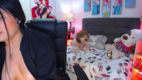 Media: Video of a cozy bedroom with a woman wearing headphones, lying on a bed with teddy bears, a black leather chair, and colorful artwork on the wall.