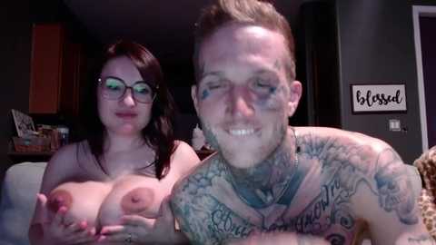 Media: Video of a pale, tattooed man with a short beard, and a fair-skinned woman with glasses, both topless, smiling in a dimly lit living room.