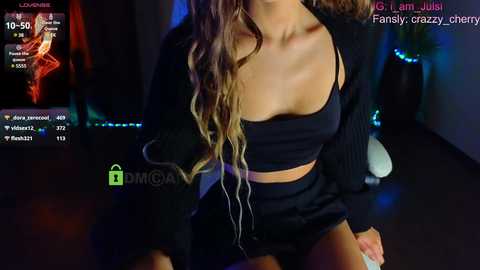 Media: Video of a young woman with long, wavy blonde hair, wearing a black crop top and shorts, sitting on the floor, dimly lit room with a \"DMG\" watermark, chat screen in the corner.