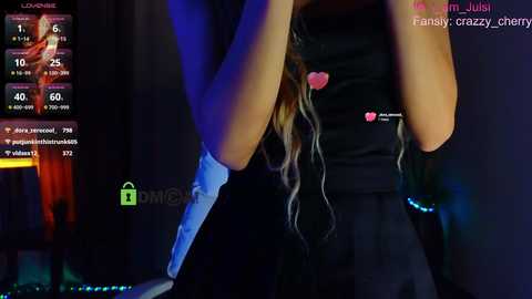 Media: Video of a young woman with long, wavy hair wearing a black shirt and a heart-shaped pendant, standing in a dimly lit room with a digital display showing social media metrics.