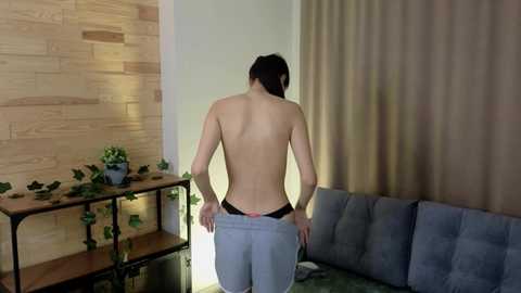 Media: Video of a topless Asian woman with short dark hair, wearing blue jeans, standing in a modern living room with wooden walls, green plants on a shelf, and beige curtains.