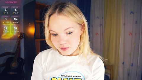 Media: Video of a young blonde woman with light skin and straight hair, wearing a white t-shirt with a logo, in a dimly lit bedroom.