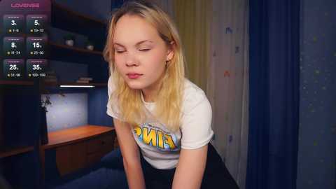 Media: A video of a blonde Caucasian teenage girl with closed eyes, wearing a \"Hanna\" t-shirt, sitting in a dimly lit bedroom with blue curtains and wooden furniture.