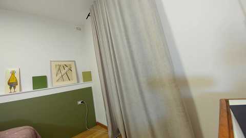 Media: A video of a minimalist bedroom with beige curtains, green and yellow framed abstract art on a white wall, a light wooden floor, and a partially visible bed.