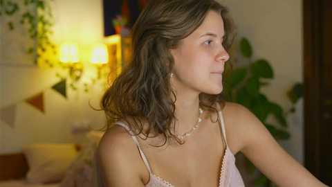 Media: Video of a young Caucasian woman with wavy, shoulder-length brown hair, wearing a light pink camisole, sitting indoors. Background features a warm, softly lit room with hanging plants and abstract art on the wall.