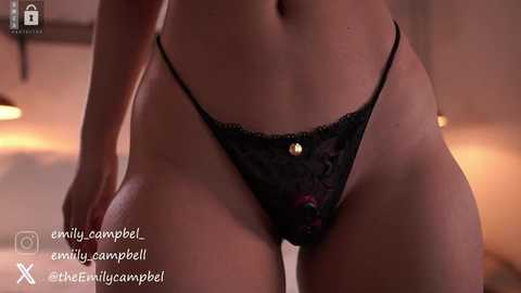 Media: Video of a woman's lower torso in black lace panties with a gold clasp, standing in a dimly lit bedroom with blurred background.