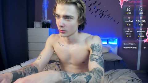 Media: A topless, young, slender, pale-skinned man with short blonde hair and numerous tattoos, sitting on a bed in a dimly lit room. The room has a white dresser, blue lighting, and a video feed overlay.