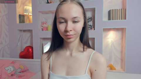 Media: A video of a young, slender, Caucasian woman with straight, light brown hair and light skin, wearing a white spaghetti-strap tank top. She stands in a softly lit room with pastel-colored shelves and a heart-shaped pillow.