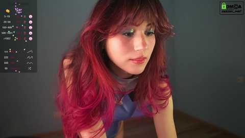 Media: Video of a young Asian woman with wavy, vibrant red hair, wearing a blue sleeveless top, looking introspective against a plain grey background.