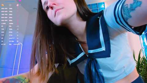 Media: Video of a young woman with fair skin, long brown hair, and a light blue sailor-style school uniform. She has a tattoo on her left arm and is smiling. Background includes a blue wall and a plant.