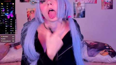 Media: Video of a young woman with blue hair, wearing a black jacket, sticking out her tongue, in a bedroom with posters and a calendar.