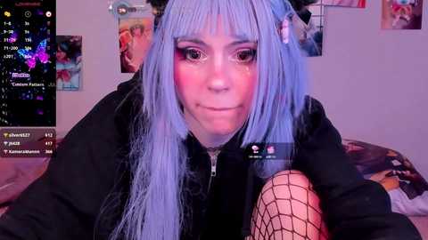 Media: Video of a young woman with long, pastel blue hair, wearing a black hoodie and fishnet tights, in a dimly lit room with posters and a gaming monitor in the background.