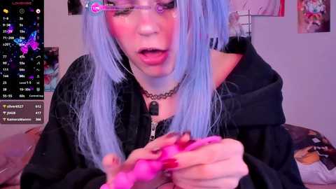 Media: Video of a light-skinned woman with blue hair, wearing glasses, a black choker, and black hoodie, playing with a pink vibrator, surrounded by colorful wall art and a digital clock.