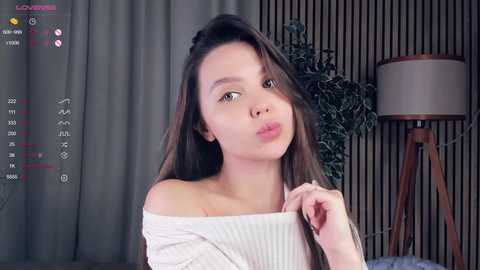 Media: A video of a young Asian woman with long brown hair, fair skin, and a small frame, wearing a white off-shoulder sweater. She's indoors with a modern decor background featuring a tall plant and a lamp.