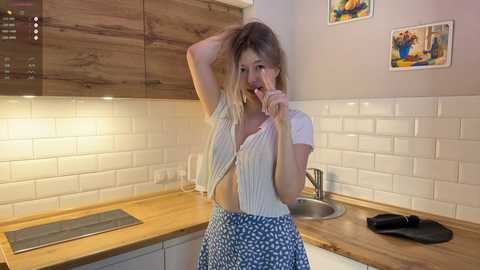 Media: Video of a young Caucasian woman with blonde hair, wearing a white cardigan and blue skirt, playfully biting her finger in a modern, white-tiled kitchen with wooden cabinets.