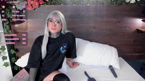 Media: A video of a young woman with long silver hair, wearing glasses, a black T-shirt, and thigh-high socks, sitting on a bed in a modern, dimly lit bedroom with wooden paneling.