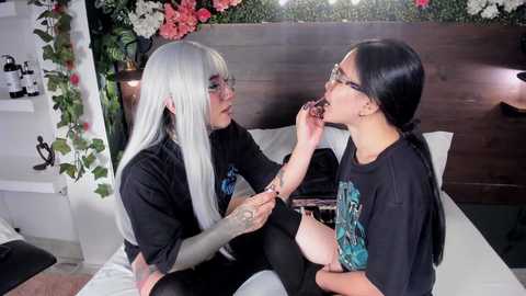 Media: Video of two women in a bedroom: one with long white hair, glasses, black shirt, and tattoos, applying makeup to another with long black hair and glasses. Background includes flowers, white shelves, and a wooden headboard.