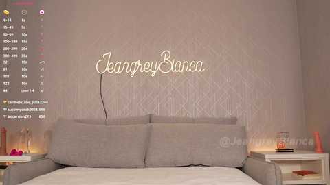 Media: Video of a modern bedroom with a beige upholstered bed against a patterned wallpaper backdrop. The name \"Jeanrey Banca\" is prominently displayed in gold cursive. The room features minimalistic decor with a nightstand and a pink candle on each side.