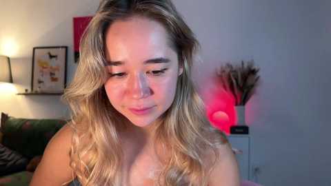 Media: Video of a fair-skinned young woman with long, wavy blonde hair, wearing a pink top, sitting indoors against a white wall, surrounded by a green couch, framed art, and a red lamp.